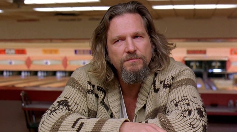 A bearded man wearing a ratty cardigan sits at a bar in a bowling alley. 