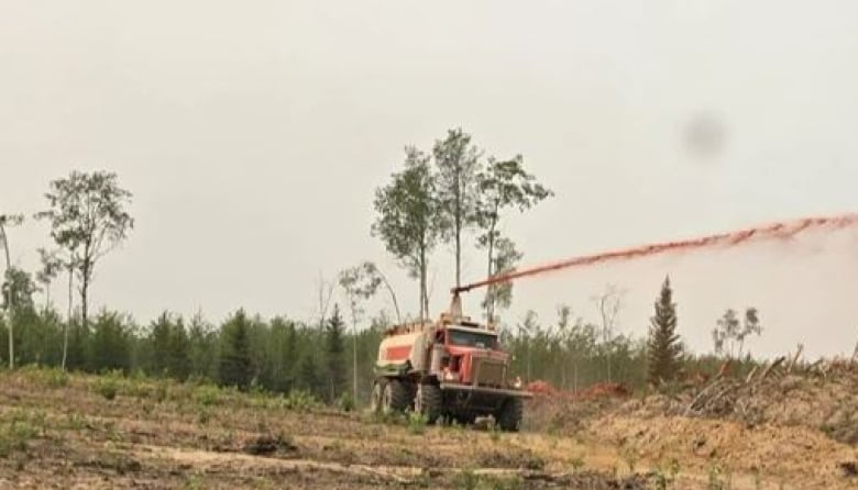 A truck sprays red liquid from a spout over grass and a pile of wood logs.