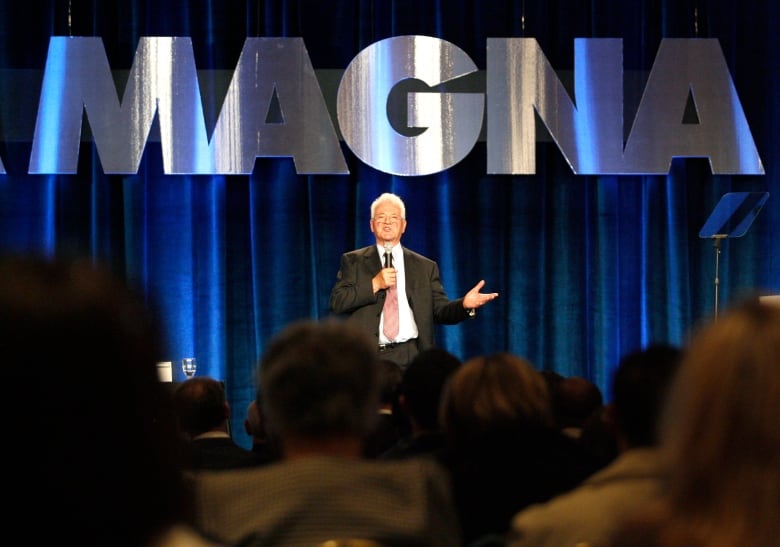 A man in a suit and tie speaks while holding a microphone below a sign marked "Magna."