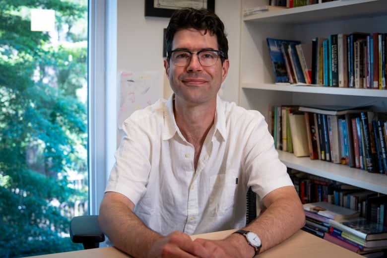 A man in a short sleeve shirt smiles as he sits in an indoor room, a bookshelf and window behind him.