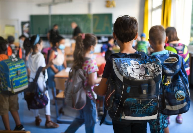 Young students with backpacks mill about as they find their seats in a brightly sunlit classroom, with teachers in blurred focus by the blackboard in the background.