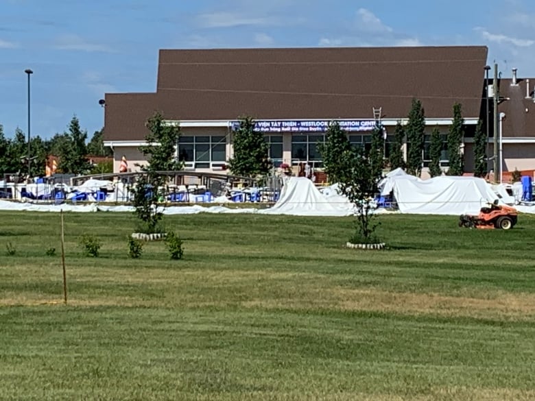 The remains of a white plastic event tent are draped onto the ground outside a building with a brown, sloped roof and a blue sign that says "Westlock Meditation Centre," as well as the centre's Vietnamese name. It is daytime, the sky is blue with a few clouds and the grass is green.