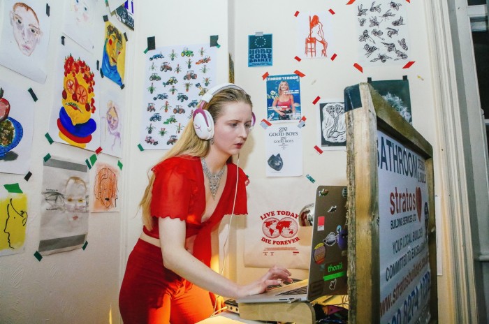 A woman in a red outfit wearing  large headphones and working on a laptop covered with stickers. The walls around her are adorned with drawings, sketches, and posters