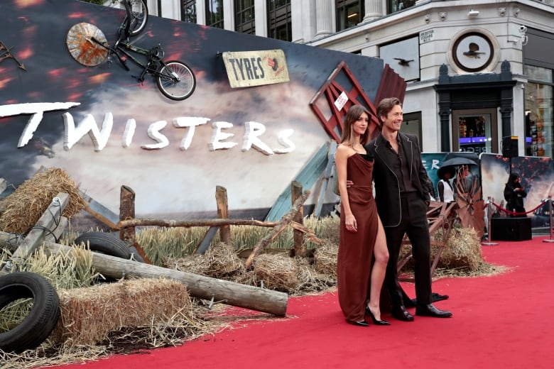 A man and woman stand on a red carpet in front of a large sign for the movie Twisters.