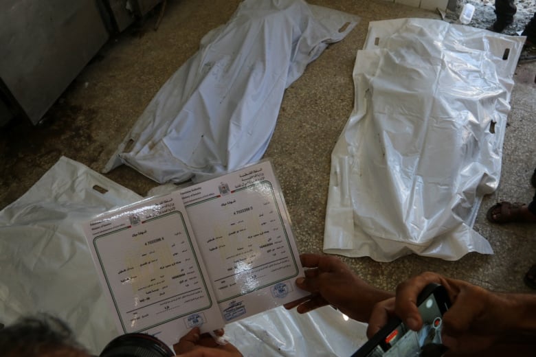 Two birth certificates are held up in front of white body bags