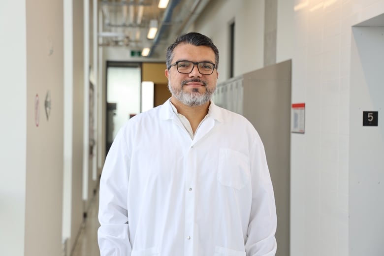 A man wearing black glasses and a white button-down shirt smiles at the camera. He is standing in a hallway near lockers and doors.