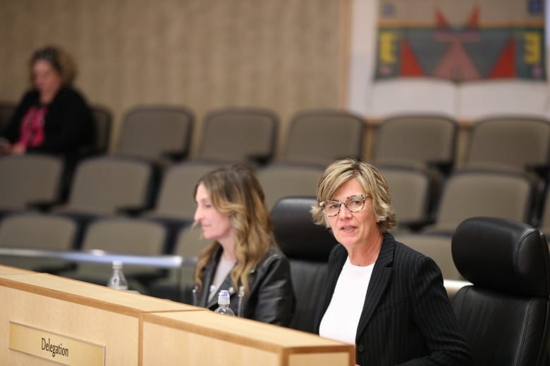 Two women, both wearing black jackets, speak while sitting at a wooden desk.