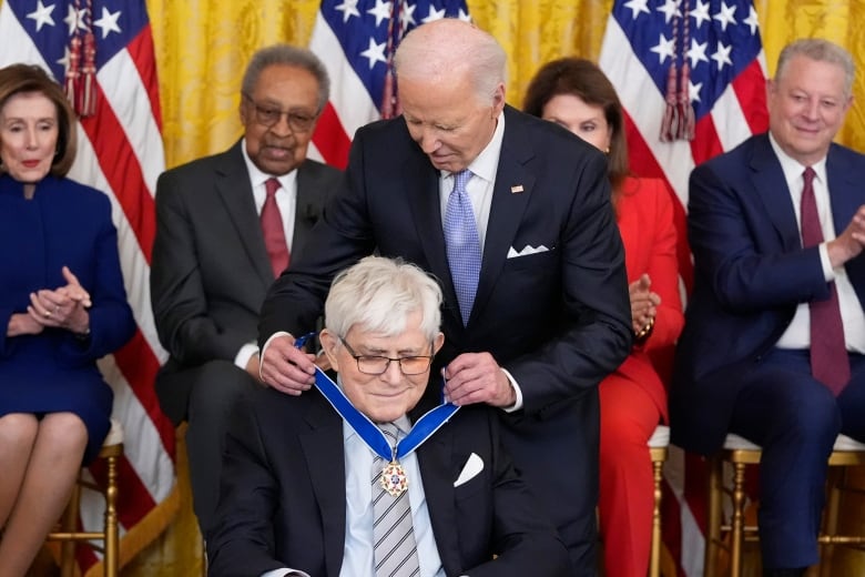 An older man stands and fastens a medal around the neck of another man seated in front of him.