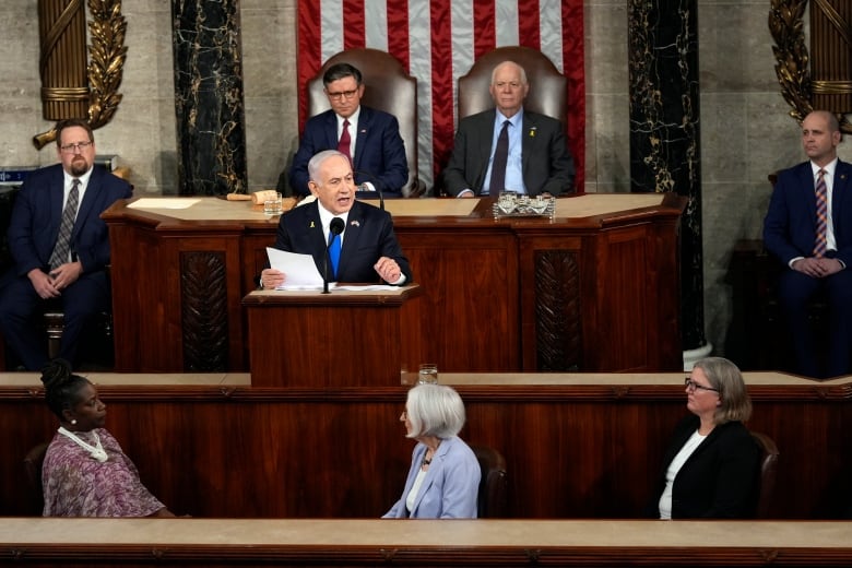 A man in a dark suit speaks while seated at a lectern in a large room decorated with an American flag as others listen. 