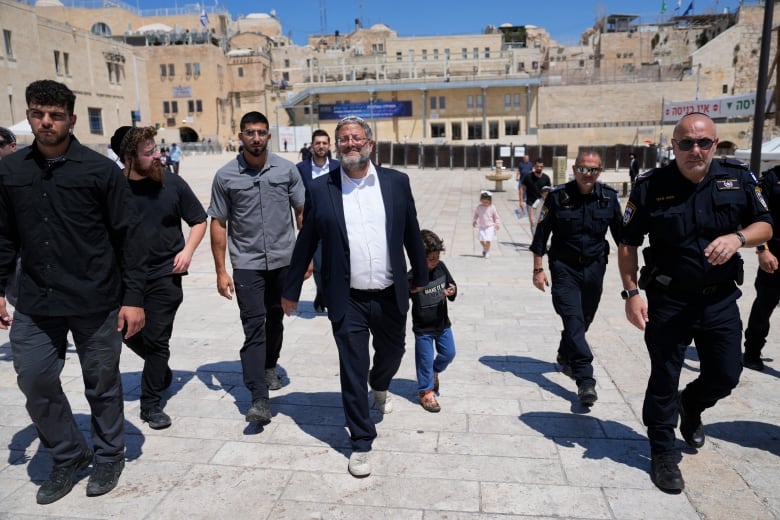 A man in a suit and white shirt flanked by several other men, walks along a stone pathway. 