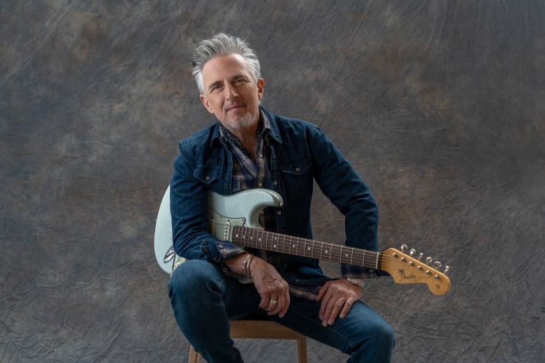 Colin James sits on a chair, holding a blue guitar.