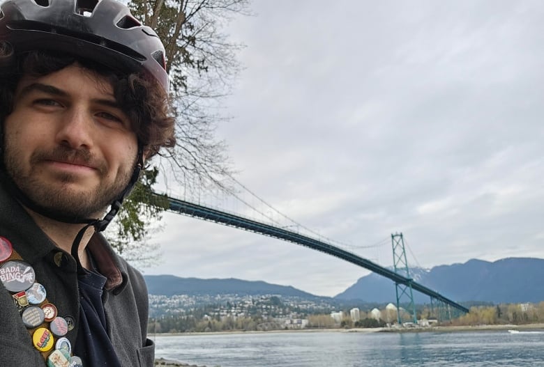 A man wearing a black bike helmet, a dark jacket and a strap covered in several pins and buttons, stands by the water with a long, green suspension bridge in the background.