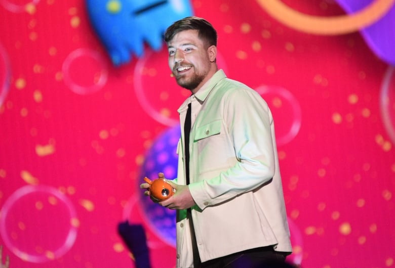 A smiling man stands in front of a pink background and holds a small award in his hands. 