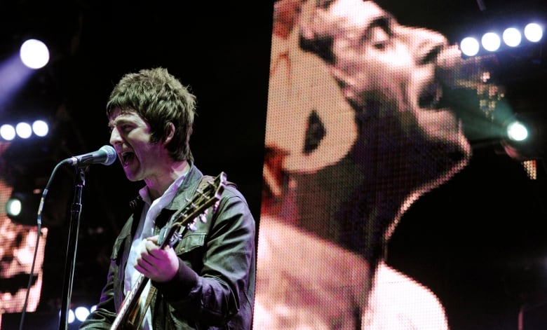 A man holding a guitar sings into a microphone in front of a large screen showing a blown up image of another man singing into a microphone. 