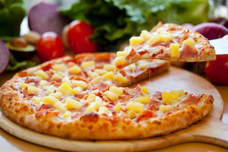 A hand lifts a size of pineapple pizza from a pie.