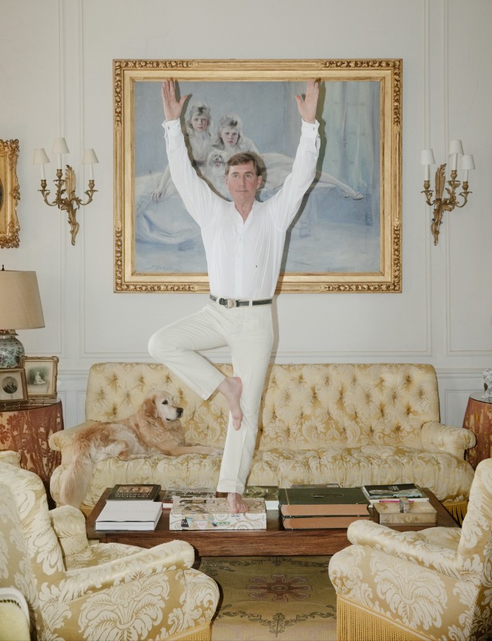 A barefoot man in white shirt and cream trousers standing on one leg on a coffee table in an ornate living room with his arms in the air