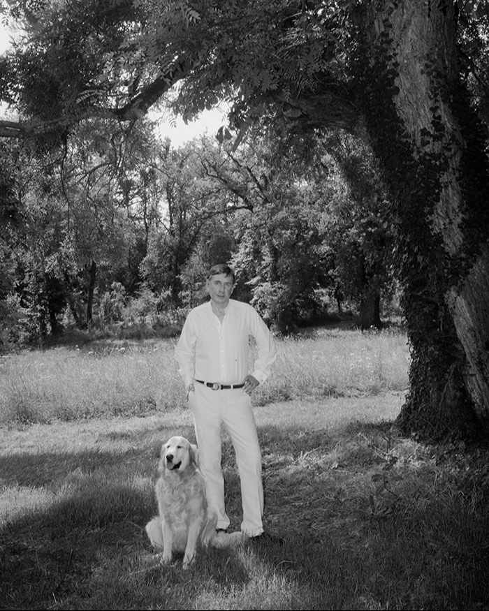 A man in white shirt and cream trousers standing under a tree in a garden with a golden retriever sitting by his side