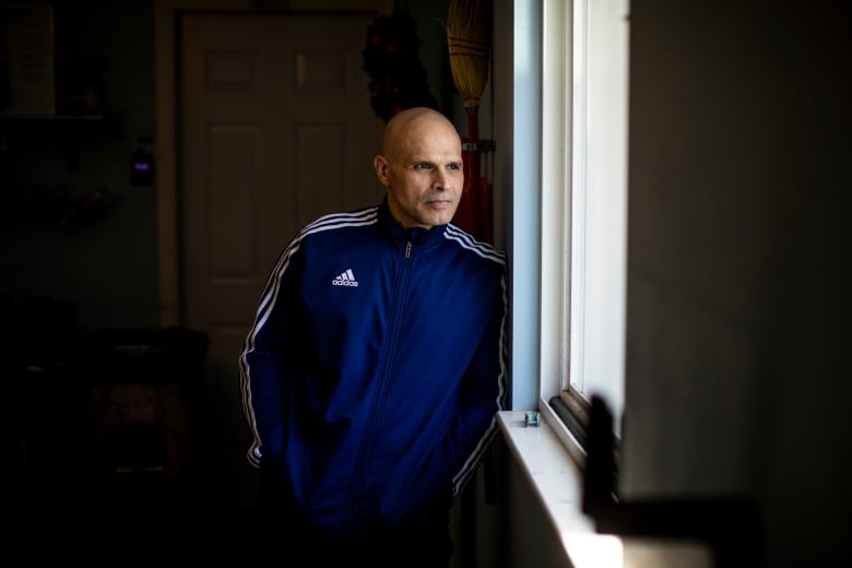 A man wearing a blue and white Adidas sports jacket standing inside a home near a window. 