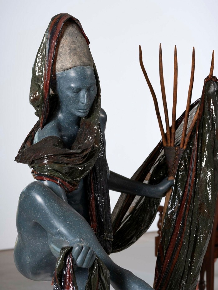 A sculpture of a woman with blue skin wearing a dark gleaming sari and clutching her raised leg in one hand 