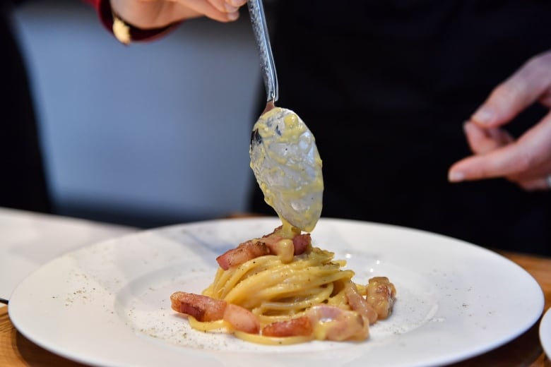 A hand spoons a yellow sauce on a plate of noodles and diced fatty pork.