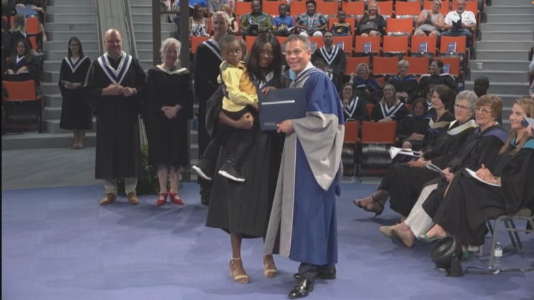 A woman in a black graduation gown holds a young child in her arms while posing for a photo. A man in a blue gown stands beside her, presenting her with a blue folder containing a diploma. 