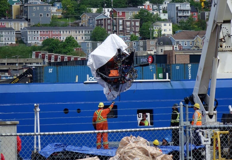 A man in orange coveralls guides wreckage being hoisted with a crane from a blue ship.