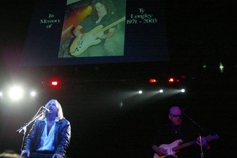 A person is seen sitting and singing onstage in a band performance, with a projection overhead displaying a guitar player.