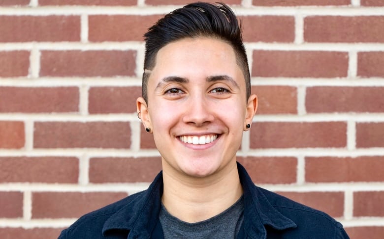 A portrait of a smiling person, wearing a grey t-shirt under dark blue denim shirt, standing in front of a brick wall.