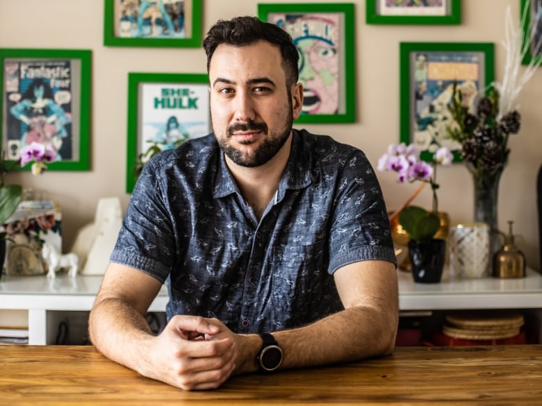 Aaron Hagey-MacKay is seated at a wooden desk, looking at the camera. Framed comic book covers are shown behind him.