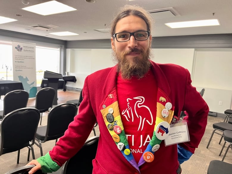 A man with a long beard smiling in a red shirt and jacket with a rainbow lapel covered in buttons. 