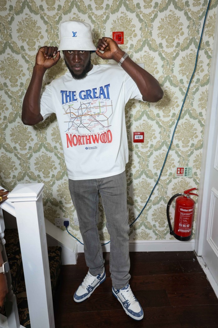 The image shows a man in white t-shirt, jeans and sneakers. He’s posing indoors, holding the brim of his hat with both hands. Behind him is a wall with vintage, floral-patterned wallpaper