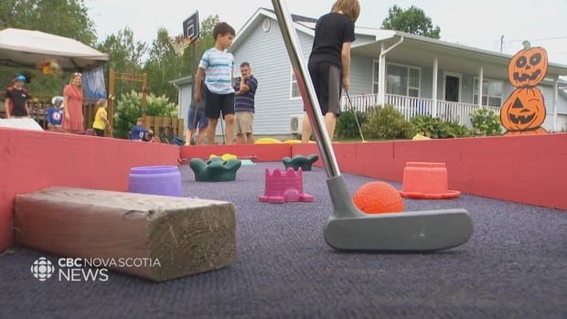 Children play mini-golf on a handmade course in a driveway while an adult looks on