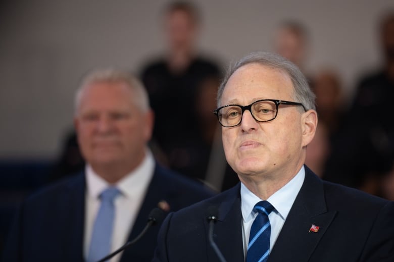 A man in dark glasses and a dark suit and tie is seen speaking at a microphone, with another man in a dark suit behind him