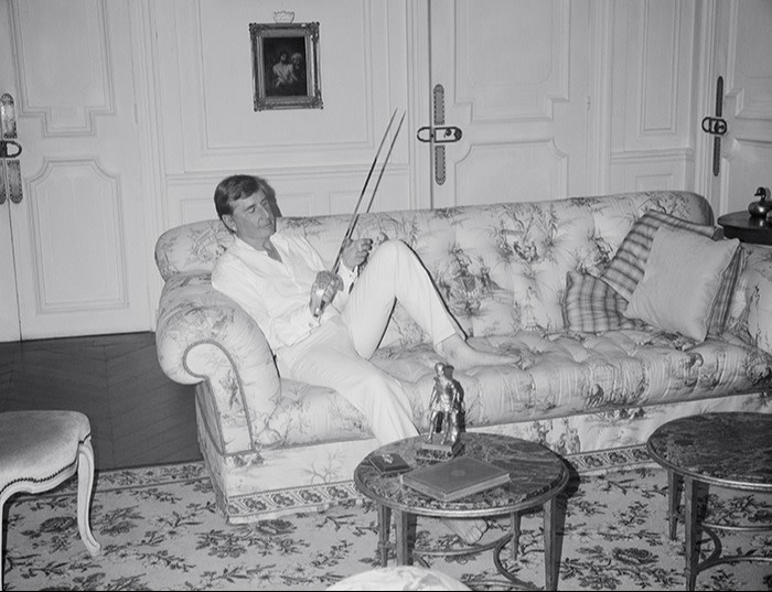 A barefoot man in white shirt and cream trousers sitting on a sofa in an ornate living room holding a fencing sword