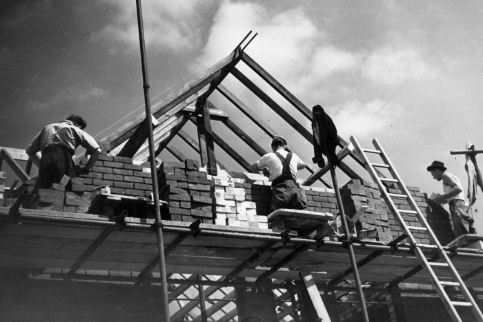 Three bricklayers work on the first storey of the building, while bare roof joists rise above them