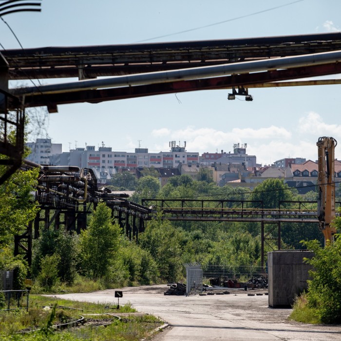 Residential buildings in the distance are visible beyond the industrial infrastructure of the Poldi area in Kladno, Czech Republic. The foreground features old industrial pipes, greenery, and a partially obstructed concrete path