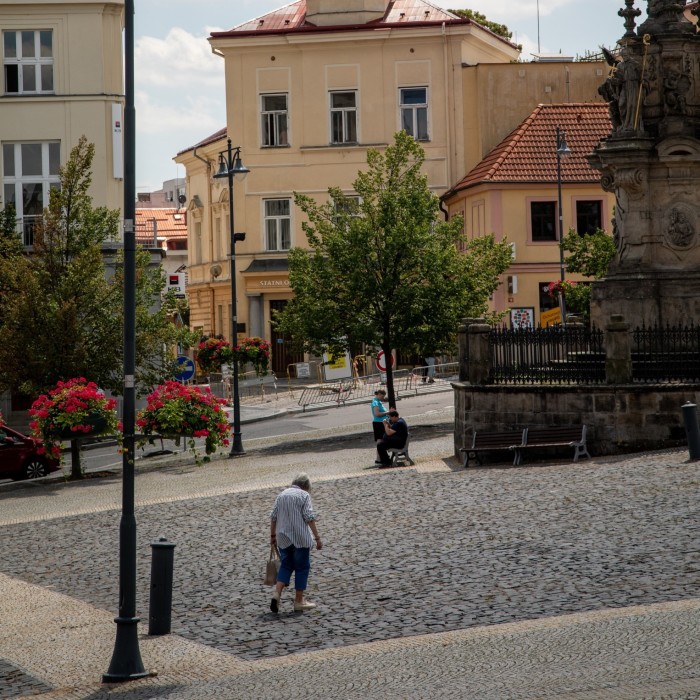 A cobblestone square in Kladno, Czech Republic, is depicted with a few people walking and sitting. The square is surrounded by historic buildings, and there are trees and flower arrangements in planters along the street. A statue or monument is partially visible on the right side of the image