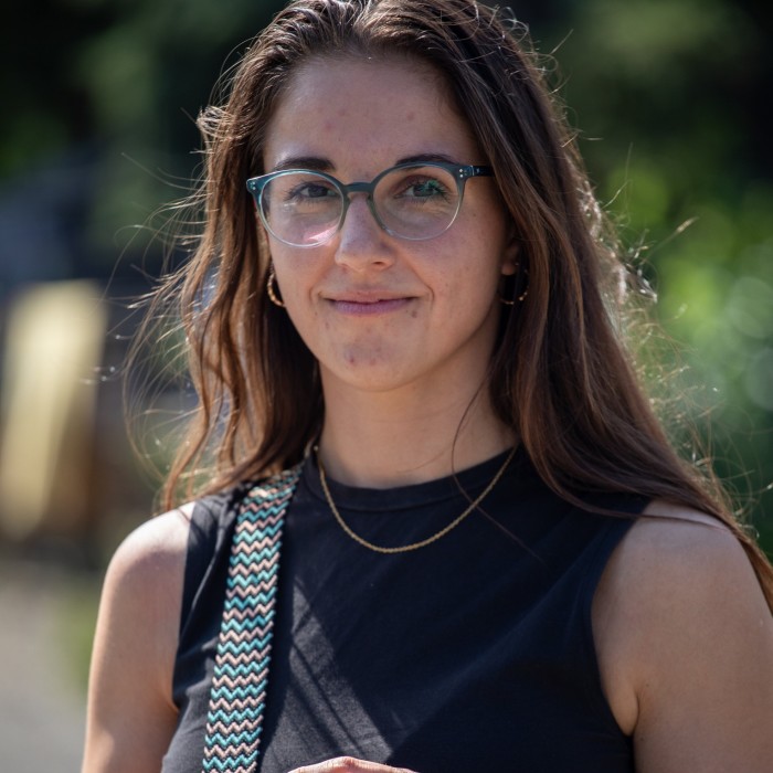 Jana Odvárková is posing for a picture in Kladno. She is wearing glasses, a black sleeveless top, and a necklace, with a patterned strap over her shoulder. The background is blurred with greenery