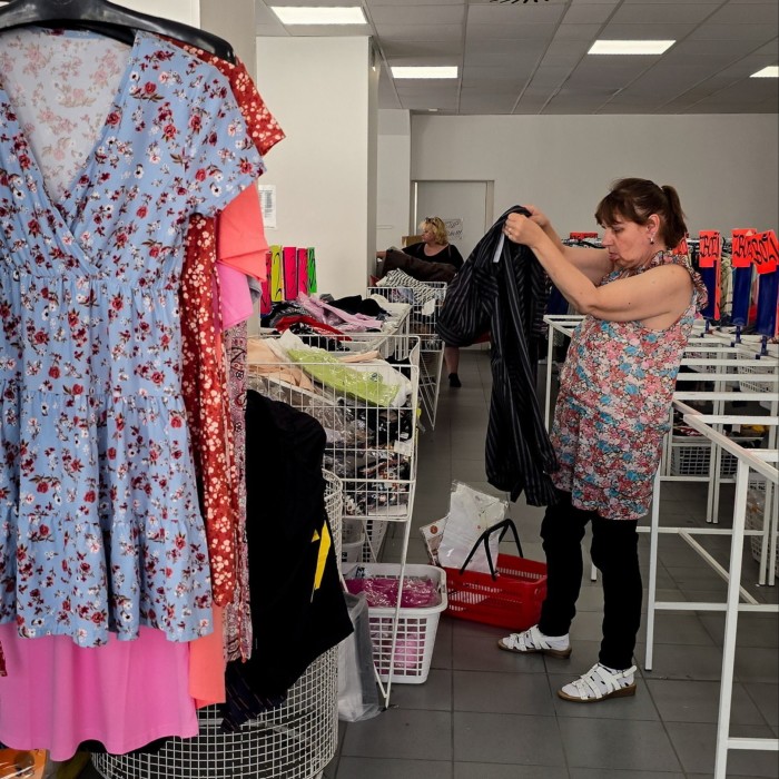 A woman in a floral dress is seen examining clothing in a discount store in Kladno. In the background, another customer is seated near bins filled with various garments. The store features racks and baskets filled with clothes, and the interior is brightly lit with fluorescent lights