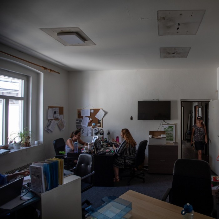 A room in the Kladno office of the People in Need. The room has several desks, with two women working at their computers and another woman walking through the doorway. The walls are adorned with bulletin boards displaying various papers and notices. A large monitor is mounted on the wall, and there are some plants on the windowsill