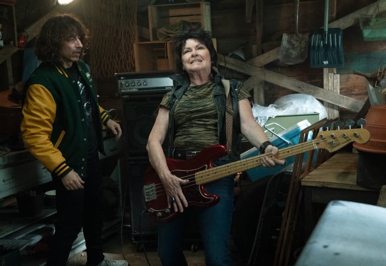 A still from a movie shows a man in a school jacket stands next to a woman holding a bass guitar in a garage.