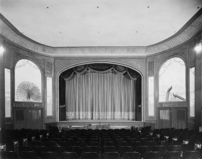 A black-and-white photograph shows a grand movie theatre with murals of peacocks on the walls. 