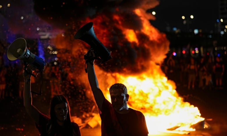 Two people hold bullhorns above their heads at night. They are backlit by a huge fire burning in the road behind them. 