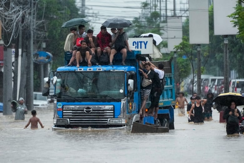 A number of people sit on top of the cab of a truck as it drives through flooded streets. Other people wade through the water next to the truck. 