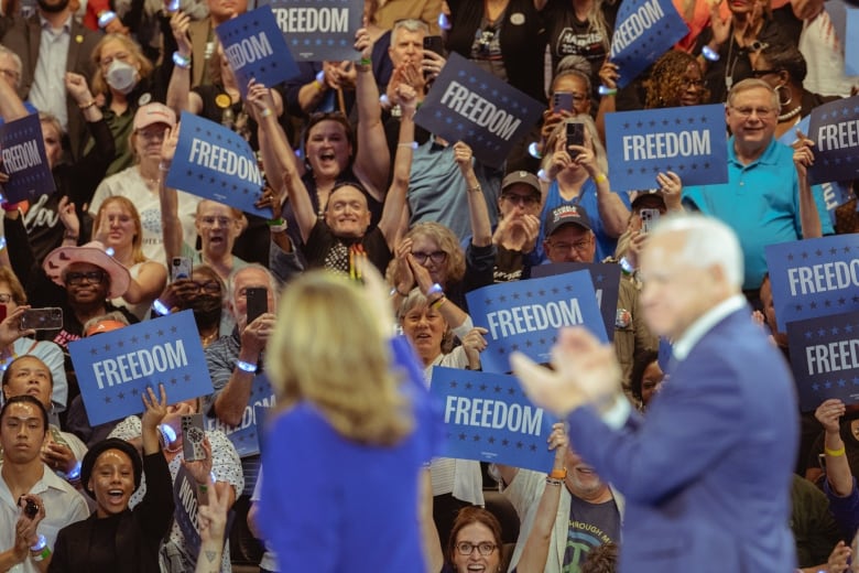 A man and a woman face a crowd of people holding signs that read 'FREEDOM' in white text on blue background. 