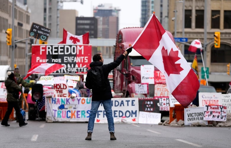 A man stands in the middle of the street waving a Canadian flag, facing a line of large trucks decked out in hand-drawn signs, including a colourful banner that reads "Thank you Truckers, Mandate Freedom" and another that says "Freedom Pour Nos Enfants"