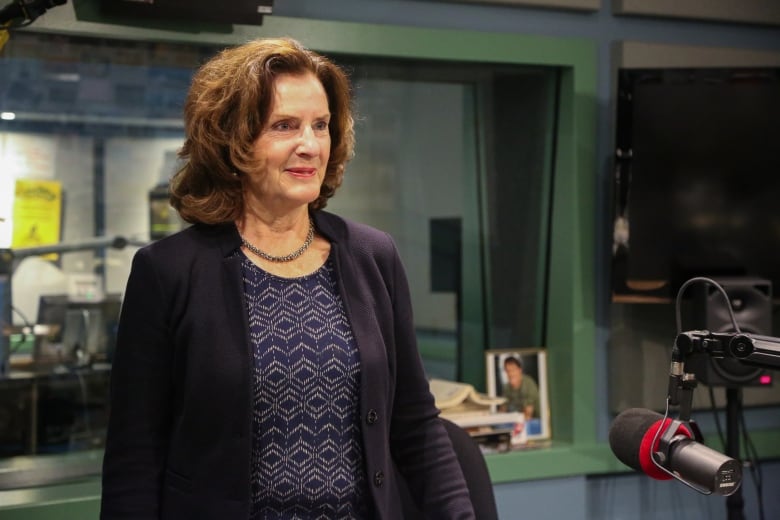 A woman with shoulder-length brown hair and a blazer stands in front of a microphone in a radio studio.