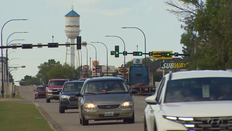 A line of vehicles drive along a street. Gas stations, fast food restaurants and other businesses are on the other side of the street. A water tower stands tall in the background.