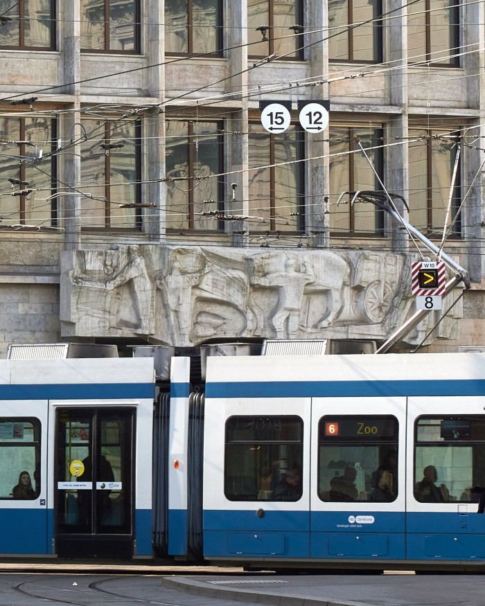 A blue and white tram passing by the Brutalist facade of Zürich’s UBS building, on the facade of which is Franz Fischer’s 1950s relief ‘Work’