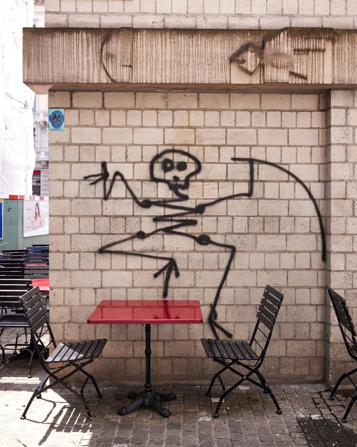 A death figure by Harald Naegeli on a grey-brick wall which appears to be dancing on a red café table in front of it